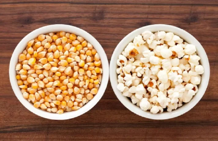 پاپ کرن و ذرت پاپ کرنی در دو کاسه سفید کنار هم روی میز قهوه ای قرار دارند.